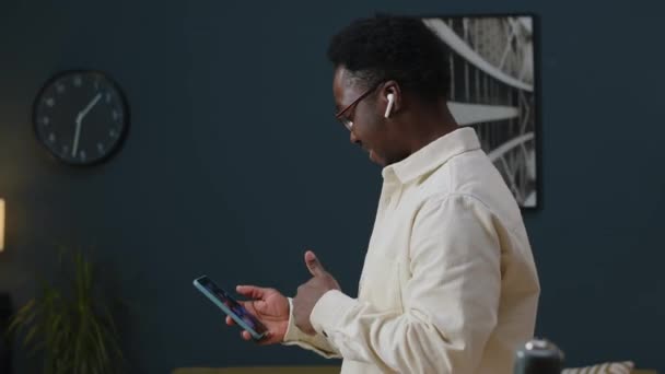 スマートフォンでビデオチャットしながら ワイヤレスイヤホンで話しながら モダンアパートに沿って歩いている若い黒人の中型ショット — ストック動画