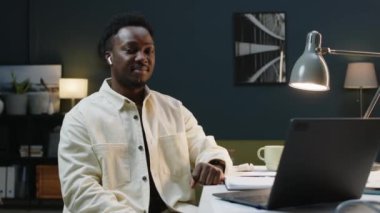 Beyaz masada, çağdaş minimalist tarzda bir ofiste otururken kameraya poz veren, kablosuz kulaklıklı genç Afro-Amerikan iş adamının orta yavaş portresi.