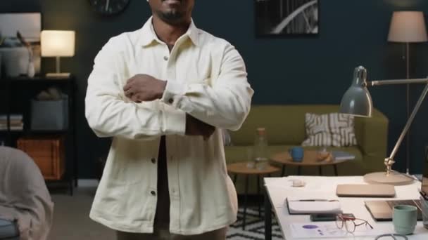 信頼できる若い黒人男性起業家のミディアムポートレートを構築し 近代的なホームオフィスに立っている耳にワイヤレスイヤホンとカメラのためのポーズ — ストック動画