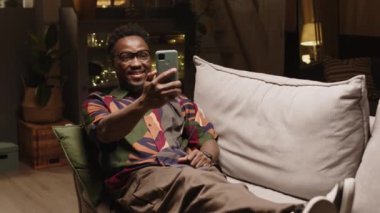 Genç hippi siyah adam rahat bir şekilde modern oturma odasındaki kanepede oturuyor. Saf sarı loş ışık ve videoyla. Akıllı telefondan arkadaşlarıyla ya da kız arkadaşıyla.