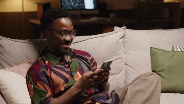 夜のコンテンポラリーアパートでソファーに座っている間 ガールフレンドとスマートフォンで明るい模様のシャツを着た若いアフリカ系アメリカ人のヒップスターのミディアムショット — ストック動画