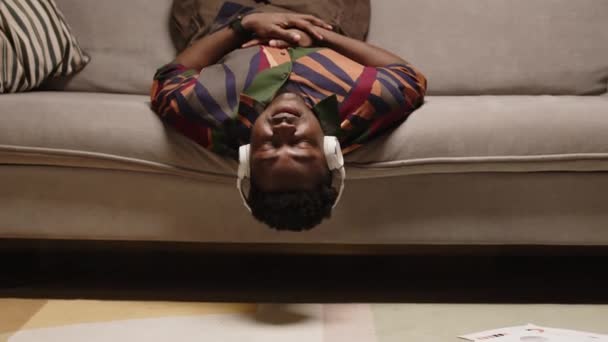 一个心平气和的非洲裔美国年轻人躺在柔软的沙发上 低着头 通过无线耳机欣赏音乐 晚上一个人在家里放松一下 — 图库视频影像