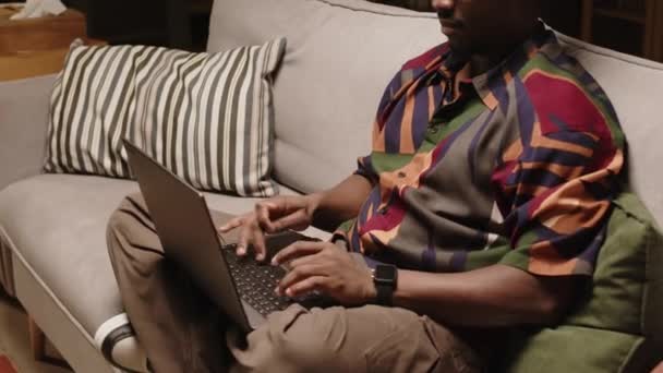 フリーランスの若い黒人男性のクローズドショット 足でソファーに座っているヒップスターの服を着て 午後自宅で働いている間にラップトップにタイピング — ストック動画