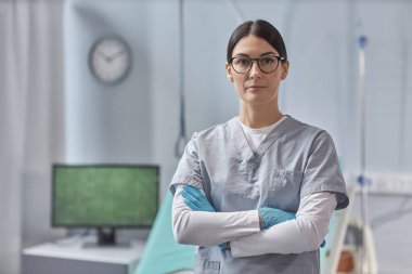 Gözlüklü ve üniformalı genç hemşire portresi kameraya bakıyor, hastane koğuşunda duruyor.