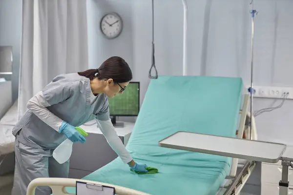 Young nurse preparing bed for patient cleaning it with sanitizer and rag in hospital ward