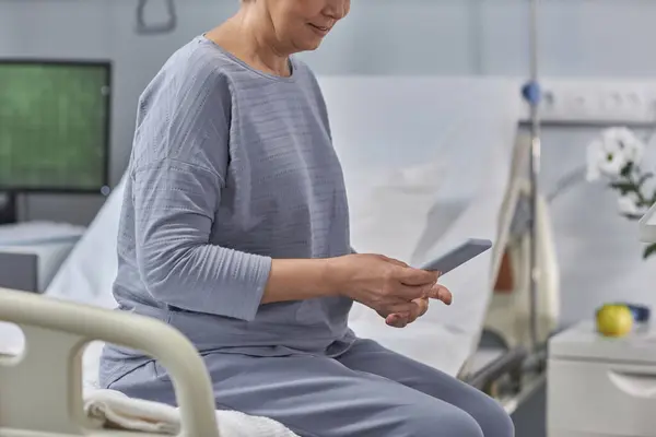Mature woman getting message on her smartphone from relatives while sitting on bed in hospital ward