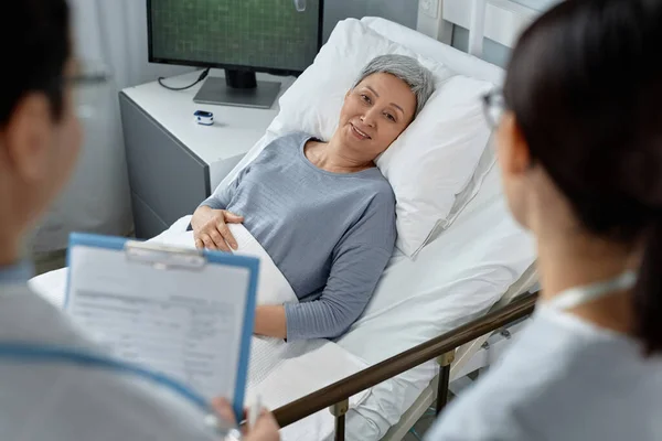 Mature woman lying on bed in ward and discussing her treatment together with doctor and nurse