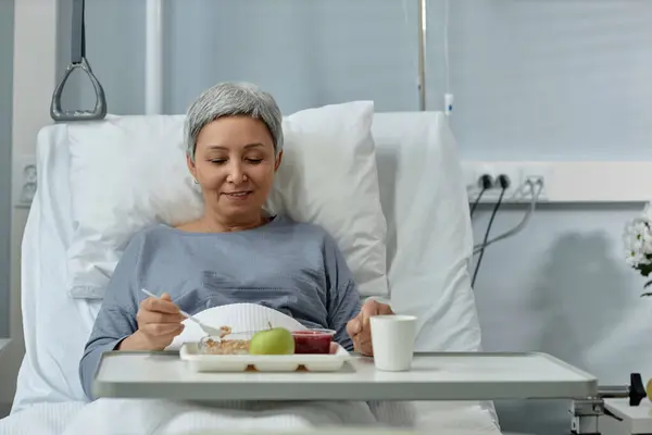 Mature woman sitting on bed and eating healthy food during lunch in ward