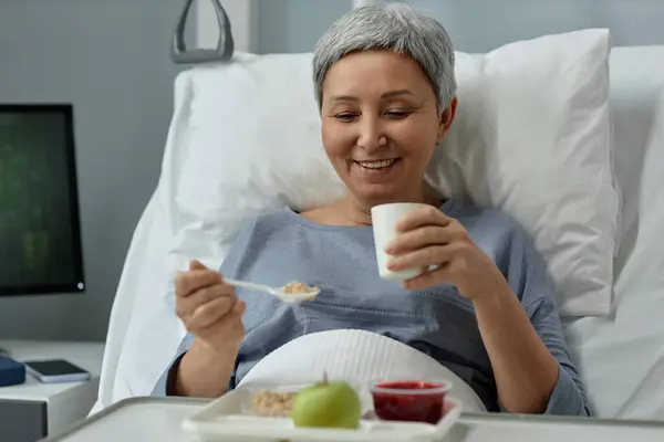 Happy mature patient sitting on bed with tray of food and enjoying her meal during her rehabilitation in hospital ward