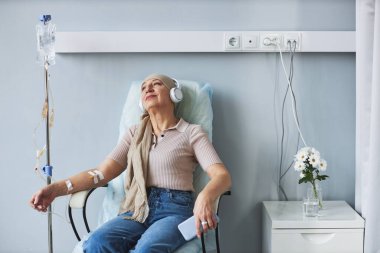 Kemoterapi sırasında serumla rahat bir sandalyede oturup müzik dinleyen son sınıf kadınının portresi.