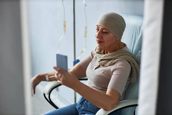 stock image Side view portrait of senior woman sitting in comfortable chair with IV drip and using smartphone