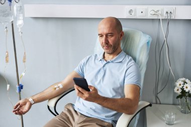 Klinikte serum damlası tedavisi sırasında akıllı telefon kullanan yetişkin bir adamın yüksek açılı portresi.