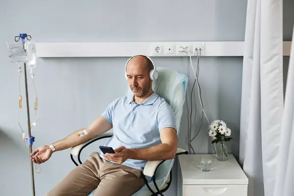 Minimal portrait of adult man using smartphone with headphones during IV drip treatment in clinic