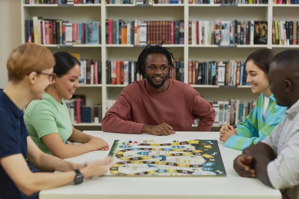 Diverse group of young people playing board games together while sitting around table in library and smiling