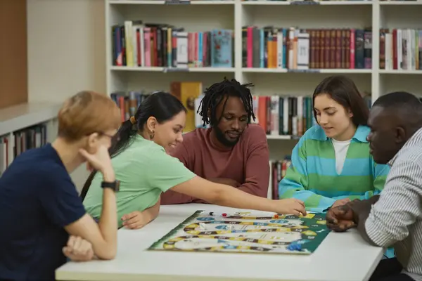 Multiethnic group of young people playing board games together while sitting around table in library and smiling