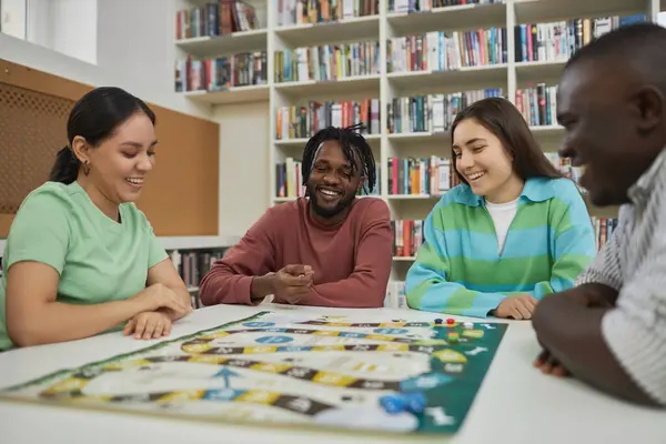 Diverse group of cheerful young people playing board games together in library and having fun