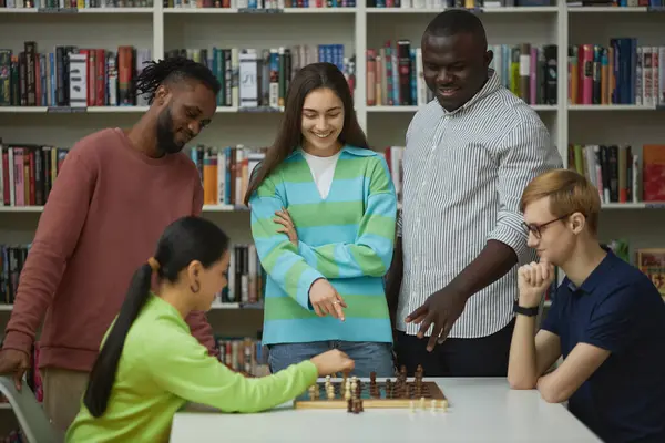 Side view at two young people playing chess with diverse group of friends watching and cheering