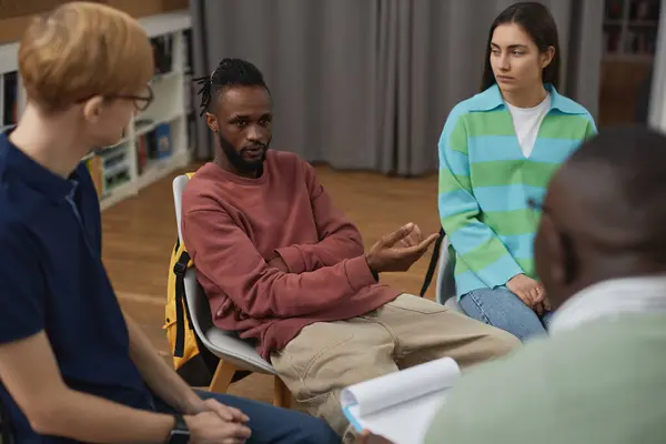 Portrait of black young man talking to psychologist during group therapy session for youth