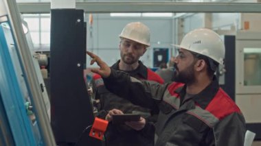 Tulumlu ve kalın şapkalı iki farklı fabrika işçisinin belini kaldırın. Makine ekipman kontrol panelini kurup çalıştırırken sohbet ediyorlar.