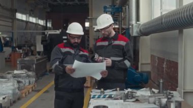 Endüstriyel fabrikada çalışırken iş kıyafetleri ve kalın şapkalı iki çok ırklı erkek teknisyenin planlarını incelerken ve tartışırken orta boy fotoğrafı.
