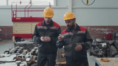 Tulumlu ve kasklı iki çok ırklı erkek çalışanın gündüz vardiyası sırasında üretim tesisinde yürürken ellerinde bulunan küçük metal makine ekipmanlarını tartışırken orta boy çekimleri.