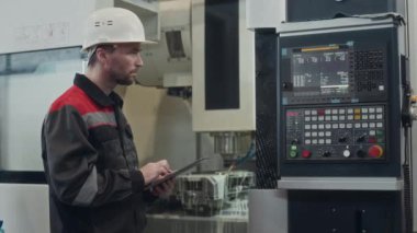 Arka planda çalışan CNC değirmen makinesini çalıştırmak için dijital tablet kullanan beyaz erkek fabrika işçisinin orta boy görüntüsü.