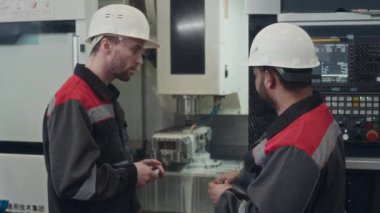 Tulumlu ve kasklı iki farklı fabrika mühendisinin küçük makine parçalarını ellerinde tutarak CNC değirmen makinesinin yanında dururken sohbet etmelerinin yan görüntüsü.