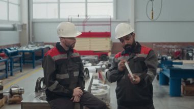 Gündüz vardiyası sırasında makinelerin metal parçalarını inceleyen çok ırklı iki erkek fabrika iş arkadaşının orta boy çekimi.
