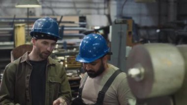 Mavi kasklı iki çok ırklı erkek çalışanın üretim tesisinde torna makinesinde çalışırken görüntüsü.