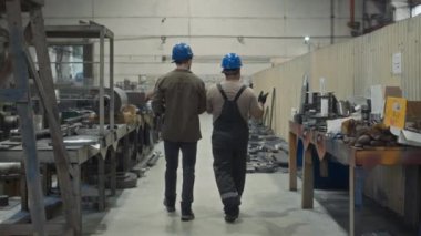 Metal fabrikasında yürüyen ve tartışan kasklı iki erkek çalışanın arka plan görüntüsü.