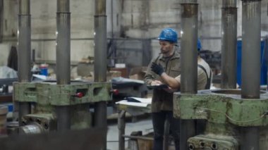 İki etnik çeşitlilikte erkek iş arkadaşının hidrolik basın makinesinin başında dikilip konuşurken, metal fabrikasında birlikte çalışırken çekilmiş görüntüleri.