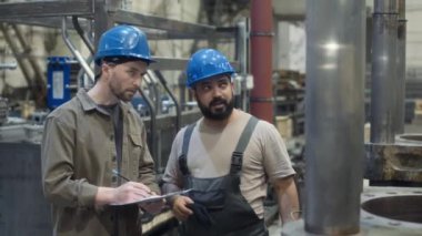 Makine ekipmanlarını kontrol ederken ve kontrol listesi üzerine notlar alırken Orta Doğulu bir erkek metal fabrikası yöneticisiyle konuşurken orta boy görüntüsü.