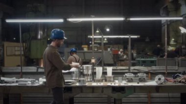Beyaz tenli erkek metal fabrikası yöneticisinin geniş bir portresi. Mavi kasketli, çalışma masasına yaslanmış, lambaların altında makine detaylarıyla kamera önünde poz veriyor.