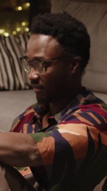 Gözlüklü genç siyahi bir adamın akşam vakti rahat bir dairede otururken kameraya baktığı dikey zoom yavaş yavaş portresi.