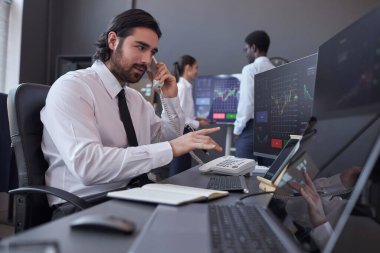 Genç komisyoncu müşteriye danışmanlık yapıyor. İş yerinde oturuyor ve bilgisayar monitörlerini çizelgelerle işaret ediyor.