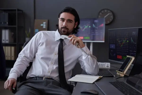 Portrait of of professional broker looking at camera while sitting with handset at his workplace