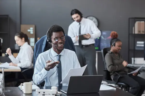 African American broker in headphones talking to customer while sitting at his workplace with laptop