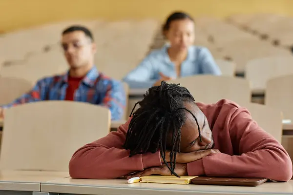 African American schoolboy lying on desk and sleeping during boring lecture with his classmates in background
