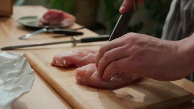 Mutfakta ahşap kesme tahtasıyla yemek pişirirken tavuk göğsünün her iki tarafını kesen tanınmamış bir erkek aşçının kırpılmış görüntüsü.