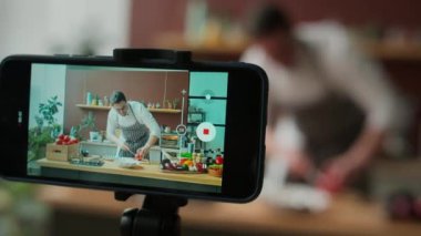 Kafkasyalı genç aşçının iş kıyafetleri içinde, mutfakta yemek videosu çekerken, ahşap tahtada greyfurtun nasıl kesileceğini gösteren akıllı telefon görüntüsüyle.