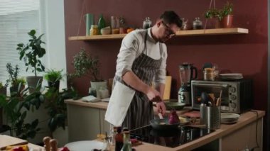 Orta boy, üniformalı, gözlüklü, beyaz bir erkek aşçının mutfak tavasında biftek pişirilirken forseps kullanması.