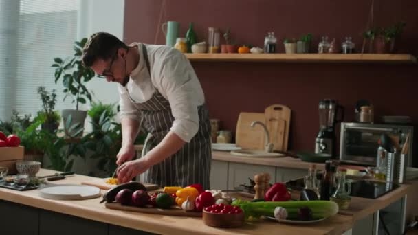明るいキッチンで調理テーブルに新鮮な野菜を切断する若い白人男性シェフのミディアムショット — ストック動画