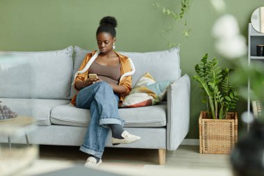 Young restful woman in casualwear with white snake on shoulders sitting on comfortable couch in living room and texting in mobile phone clipart