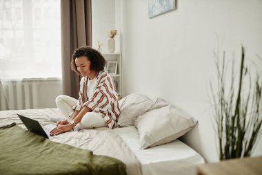 Yatakta dizüstü bilgisayar kullanan siyahi genç bir kadının yan görünüm portresi ve minimal ev içi gülümsemesi, kopyalama alanı