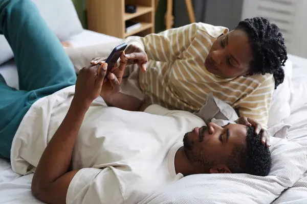 African American couple playing game on smartphone while lying on bed in bedroom