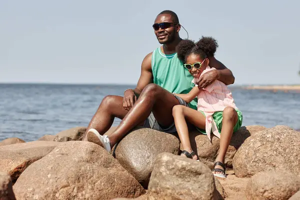 African American dad sitting on stones on coastline together with his daughter and they enjoying summer days
