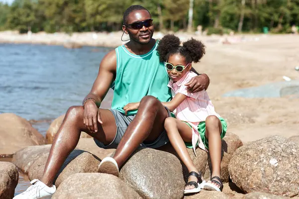 African American dad sitting on stones on the beach together with his daughter and enjoying summer vacation