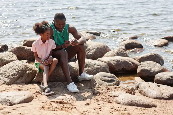 African American dad spending time with his daughter outdoors, they sitting on stones on the beach