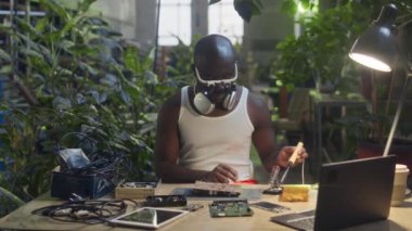 Beyaz fanila ve saç bandı takan genç siyah erkek elektronik mühendisinin beli yeşil bitkilerle dolu fabrikada anakart parçalarını lehimliyor.