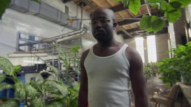 Beyaz atlet ve gözlüklü genç kaslı siyah adamın düşük açılı portresi imalat fabrikasında yeşil bitkilerin arasında kameraya bakıyor.